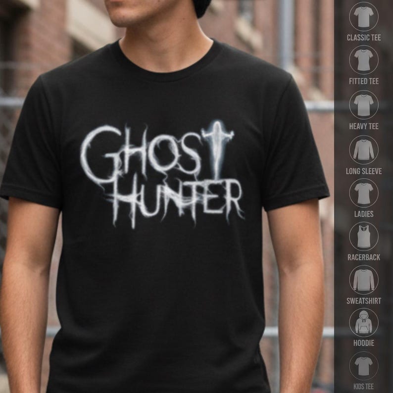 May include: A black short-sleeved t-shirt featuring the white text GHOST HUNTER in a stylized, wispy font that resembles smoke or mist. The letter T in GHOST is replaced by a small, glowing white silhouette of a figure with arms outstretched.