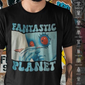 Pode incluir: Uma t-shirt preta de manga curta com uma impressão gráfica do filme Fantastic Planet. O design mostra uma personagem de pele azul com olhos vermelhos grandes segurando um pequeno objeto perto do rosto, com o texto Fantastic Planet em azul.