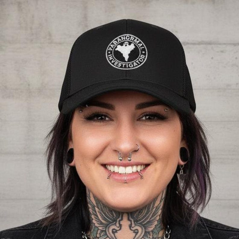 May include: Black trucker hat featuring a white embroidered circular design with the text "PARANORMAL INVESTIGATOR" and a ghost graphic. The hat has a mesh back and a curved peak.