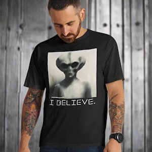 May include: A black short-sleeved t-shirt featuring a square graphic print of a grey alien with large dark eyes. Below the alien illustration, the text I BELIEVE is printed in a simple white sans-serif font. The shirt has a classic crew neckline.