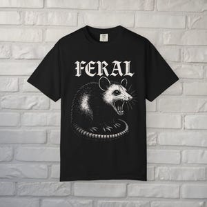 Puede incluir: Una camiseta negra de manga corta con una ilustraci&oacute;n gr&aacute;fica blanca de una zarig&uuml;eya gru&ntilde;endo. Sobre la zarig&uuml;eya, la palabra FERAL est&aacute; impresa en una fuente blanca, negrita y estilizada. La camiseta tiene cuello redondo y corte recto.