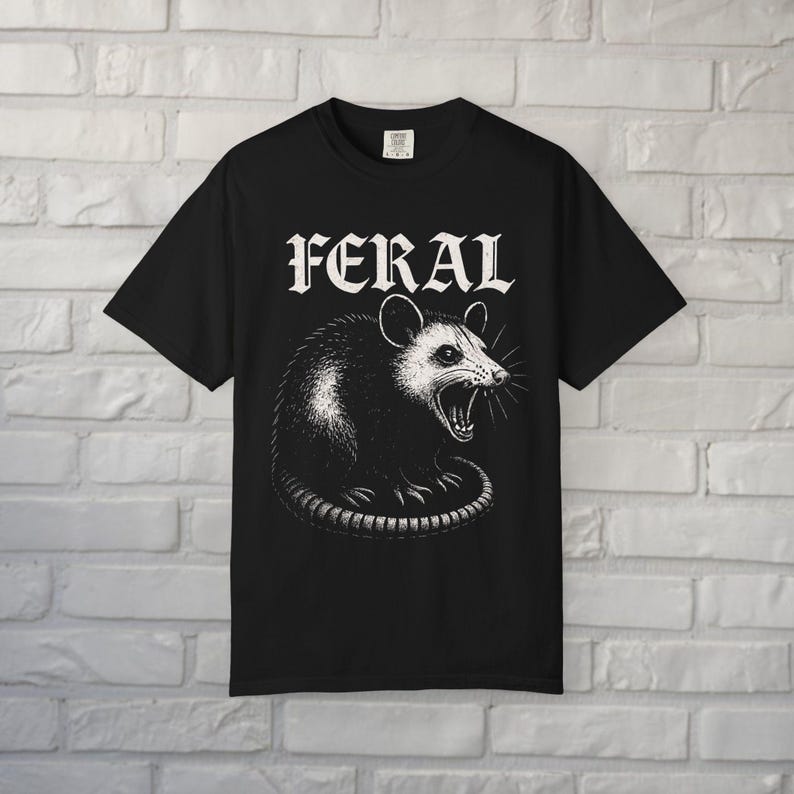 Puede incluir: Una camiseta negra de manga corta con una ilustraci&oacute;n gr&aacute;fica blanca de una zarig&uuml;eya gru&ntilde;endo. Sobre la zarig&uuml;eya, la palabra FERAL est&aacute; impresa en una fuente blanca, negrita y estilizada. La camiseta tiene cuello redondo y corte recto.