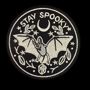 May include: A black and white embroidered patch with a bat, crescent moon, stars, and leaves. The text "STAY SPOOKY" is stitched around the edge.