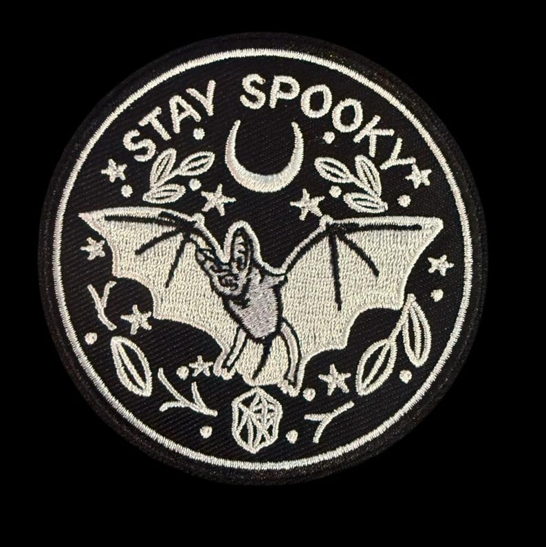 May include: A black and white embroidered patch with a bat, crescent moon, stars, and leaves. The text "STAY SPOOKY" is stitched around the edge.