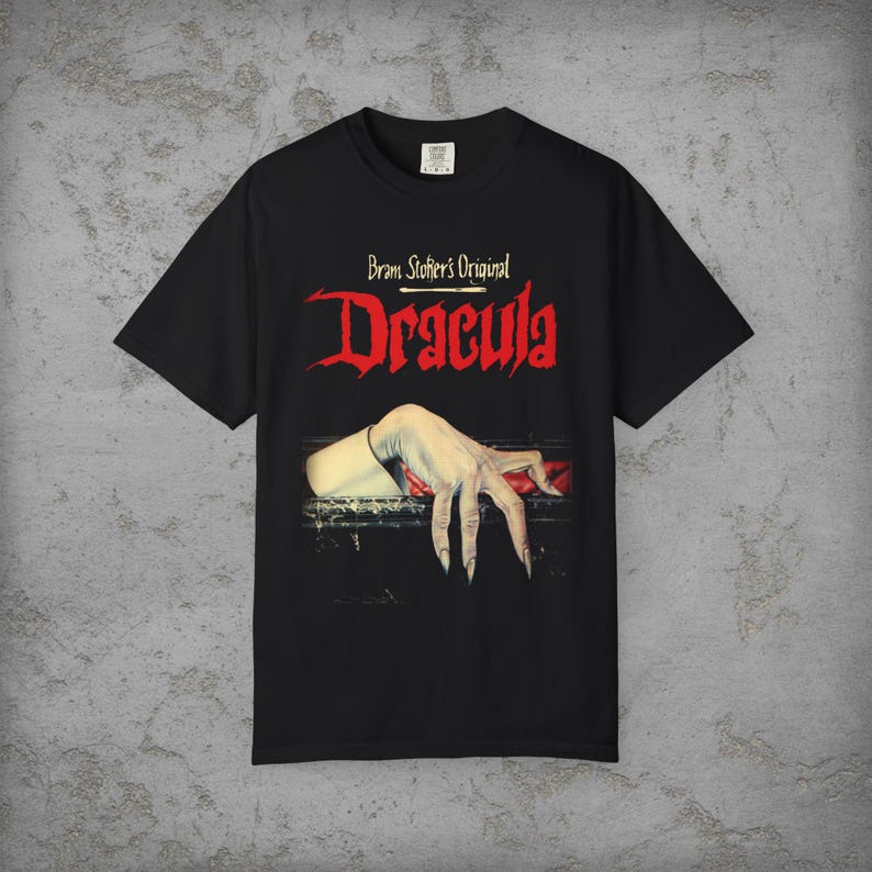 May include: A black short-sleeve t-shirt featuring the text Bram Stoker's Original Dracula in red and white lettering. The graphic depicts a pale, clawed hand emerging from a dark, rectangular opening with a red fabric accent underneath.