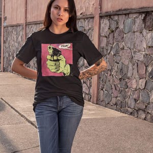 A young woman wearing a black t-shirt with a graphic of a gun and the word "PRIZE" printed on it, standing in front of a stone wall.