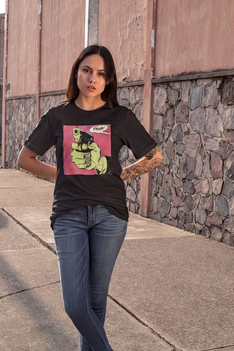 A young woman wearing a black t-shirt with a graphic of a gun and the word "PRIZE" printed on it, standing in front of a stone wall.