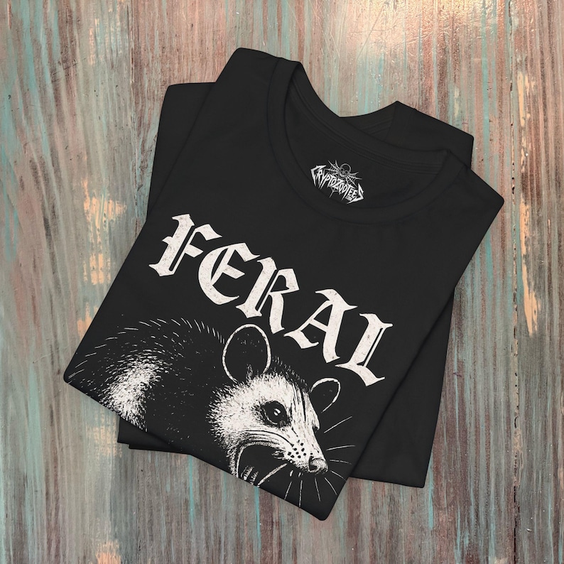 Puede incluir: Una camiseta negra con un gr&aacute;fico blanco de una zarig&uuml;eya con la boca abierta. La palabra FERAL est&aacute; impresa en una fuente blanca desgastada sobre el animal. La etiqueta interior del cuello muestra el texto Cryptozootees con un icono de calavera.