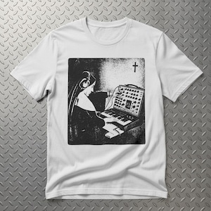 Könnte beinhalten: Ein weißes kurzärmeliges T-Shirt mit einem schwarz-weißen Grafikdruck, der eine Person in einer Tracht mit Kopfhörern zeigt, die einen modularen Synthesizer spielt. Ein kleines Kreuzsymbol befindet sich in der oberen rechten Ecke der Grafik.