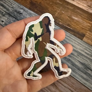 May include: A Bigfoot patch with a camouflage design in shades of green, brown, and tan. The patch has a white border and depicts the silhouette of the legendary creature. Perfect for jackets, bags, or other fabric items.