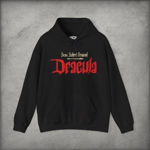 May include: A black hooded sweatshirt featuring a front graphic with the text Bram Stoker's Original in white and the word Dracula in a large, jagged red font. The garment includes a drawstring hood and a large front kangaroo pocket.