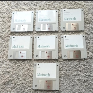 May include: Seven vintage Macintosh floppy disks. Each disk is light gray with the word "Macintosh" printed in green. Some disks have additional text, including "Install Me First", "Install 2", "Tabs", "Printing", and "Fonts". The Apple logo is visible.