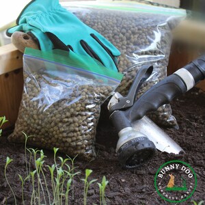 May include: Two clear plastic bags filled with brown rabbit manure pellets. A green gardening glove, a black garden hose nozzle, and a few green seedlings are in the dirt.