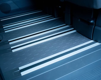 May include: A vehicle interior floor featuring dark gray rubberized matting with a repeating chevron texture. The floor is divided by parallel silver metallic tracks that run longitudinally, providing a structured surface for seating or cargo.