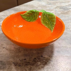 May include: A bright orange ceramic bowl shaped like a citrus fruit, featuring a textured surface that mimics the skin of an orange. Two sculpted, glossy green leaves are attached to the inner rim, adding a realistic botanical detail to the piece.