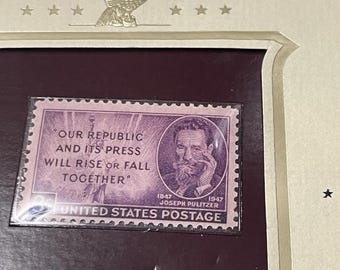 May include: A framed United States postage stamp featuring a portrait of Joseph Pulitzer and the quote "Our Republic and its Press Will Rise or Fall Together." The stamp is purple with white text and is set against a dark burgundy background.