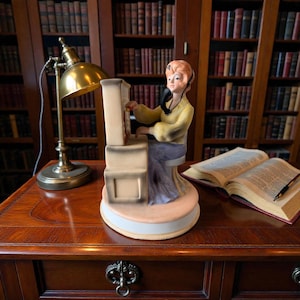 May include: A ceramic figurine of a person with orange hair wearing a yellow long-sleeved top and a light purple skirt, seated at a beige switchboard console while holding a black telephone receiver to their ear, mounted on a circular base.