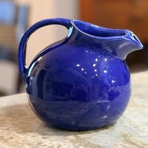 May include: A round, cobalt blue ceramic pitcher with a glossy finish, featuring a curved handle on the side and a wide, integrated pouring spout. The surface of the vessel is smooth and reflective, showing light highlights across its rounded body.