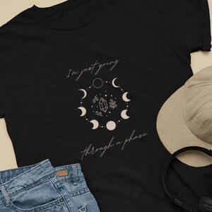 Könnte beinhalten: Schwarzes T-Shirt mit einer weißen Grafik eines Kristalls und Mondphasen. Der Text "I'm just going through a phase" ist auf dem Shirt geschrieben.