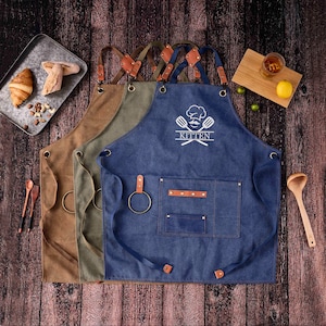 May include: Three aprons in brown, olive green, and blue denim with leather details and a metal ring. The blue apron features the word "KITTEN" and a chef's hat logo. The aprons are displayed on a wooden surface.