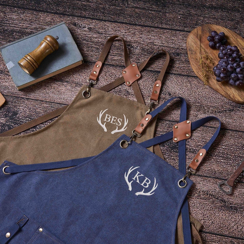 May include: Two brown and blue canvas aprons with adjustable straps and leather accents. The aprons have embroidered antlers with the initials "BES" and "KB".