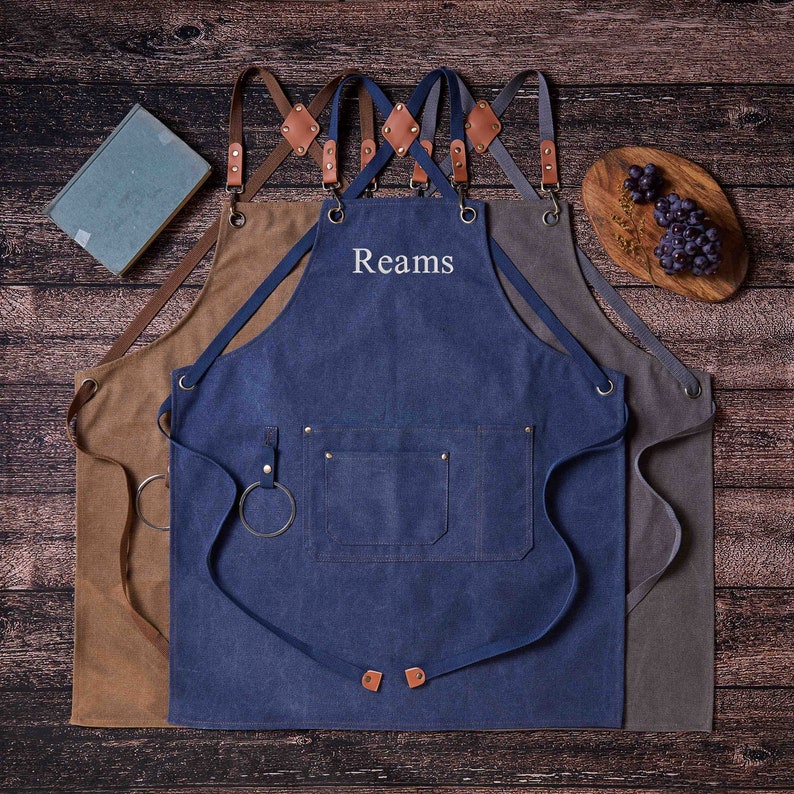 May include: Three denim aprons with brown leather straps. The center apron has the name "Reams" embroidered on it in white thread.