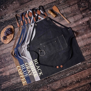 May include: A set of six denim aprons in different colors, including black, white, gray, blue, and coffee. Each apron has a large pocket and is labeled with its color.