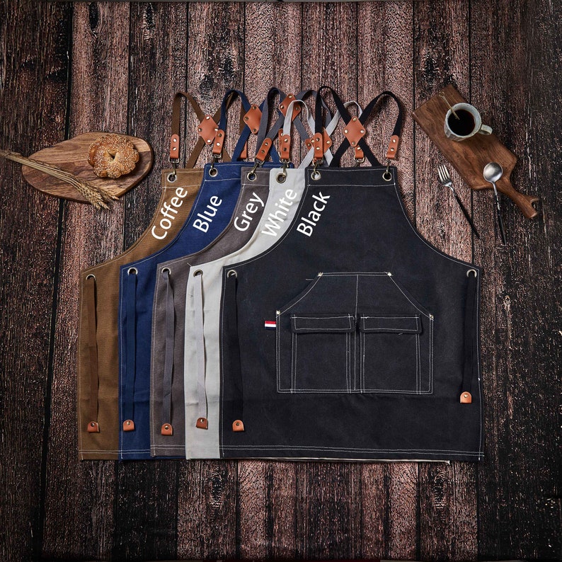 May include: A set of 6 canvas aprons in different colors, including coffee, blue, grey, white, and black. Each apron has adjustable straps and two pockets. The aprons are lined up in a row, with the straps facing the viewer.