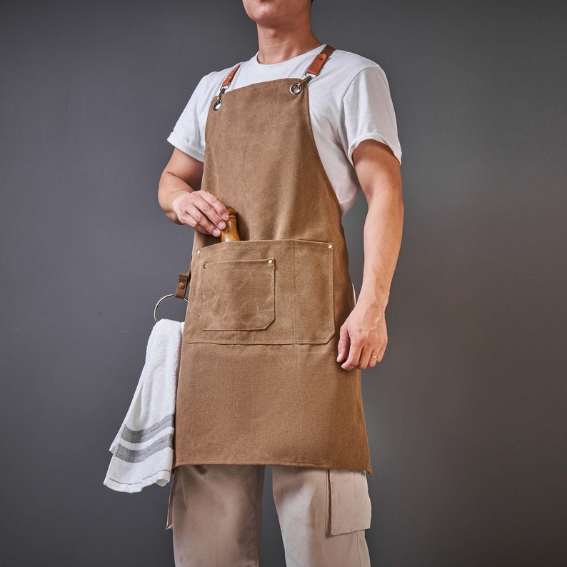 May include: A brown canvas apron with a large front pocket and adjustable straps. The apron has a leather detail on the straps and a metal ring on the left strap.