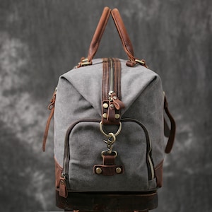 May include: A gray canvas duffel bag with brown leather accents and a zipper closure. The bag has a top handle and a detachable shoulder strap.
