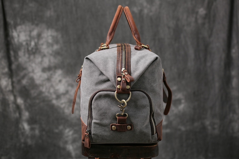 May include: A gray canvas duffel bag with brown leather accents and a zipper closure. The bag has a top handle and a detachable shoulder strap.