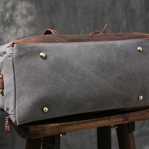 May include: A gray canvas and brown leather duffel bag with brass accents. The bag is sitting on a wooden stool.