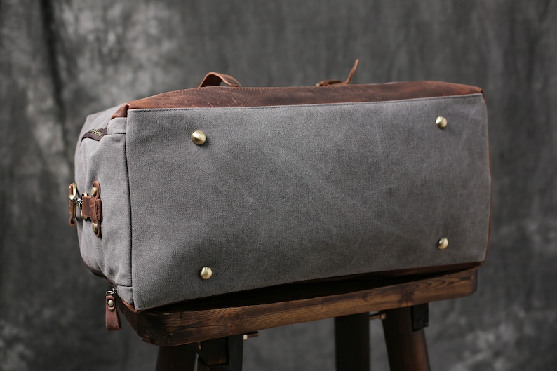May include: A gray canvas and brown leather duffel bag with brass accents. The bag is sitting on a wooden stool.