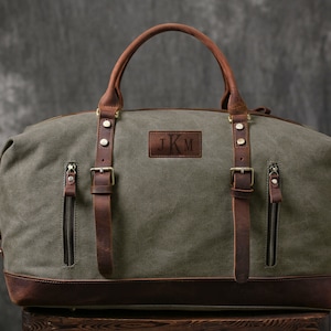 May include: A green canvas and brown leather duffel bag with a leather strap and buckle closure. The bag has a personalized leather patch with the initials "JKM".