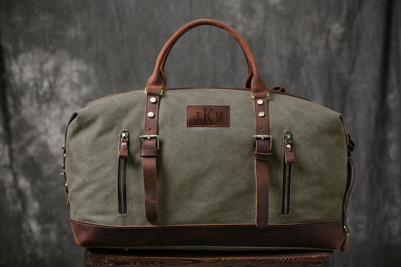 May include: A green canvas and brown leather duffel bag with a leather strap and buckle closure. The bag has a personalized leather patch with the initials "JKM".