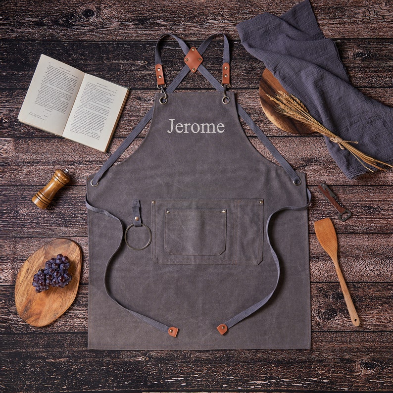 May include: A gray canvas apron with adjustable straps and a large pocket. The apron has the name "Jerome" embroidered on the front.