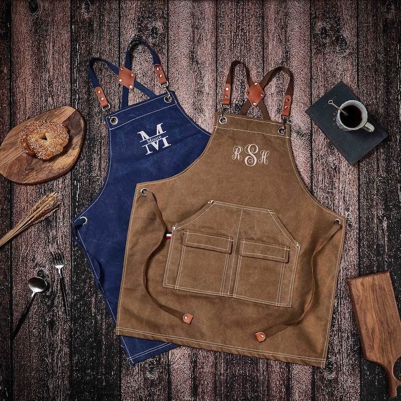 May include: Two canvas aprons with adjustable straps and pockets. One apron is brown with white stitching and the initials "RSH" embroidered on the front. The other apron is blue with white stitching and the initials "TM" embroidered on the front.