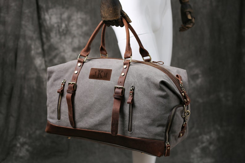 May include: A gray canvas duffel bag with brown leather accents and straps. The bag has a zipper closure and a leather patch with the initials "J.K.M." on the front.