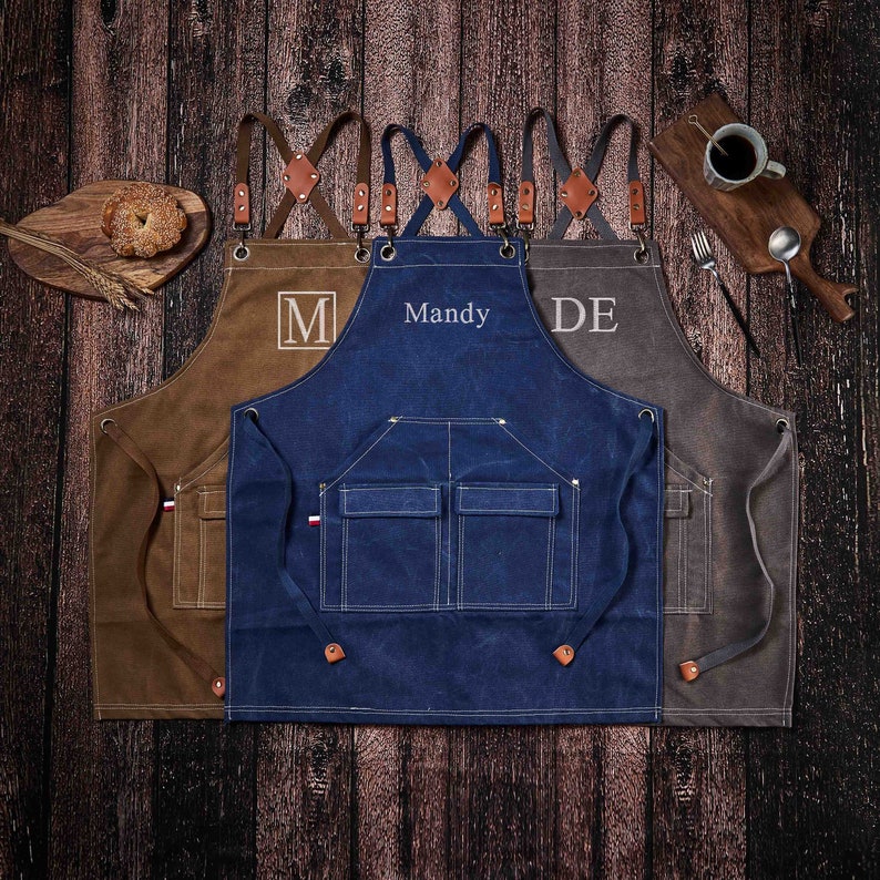 May include: Three canvas aprons with adjustable straps and leather accents. The aprons are brown, blue, and gray. The brown apron has the letter "M" embroidered on it. The blue apron has the name "Mandy" embroidered on it. The gray apron has the letters "DE" embroidered on it.