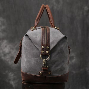 May include: A gray canvas and brown leather duffel bag with a zipper closure and a top handle. The bag has a brass buckle and a brass hook.
