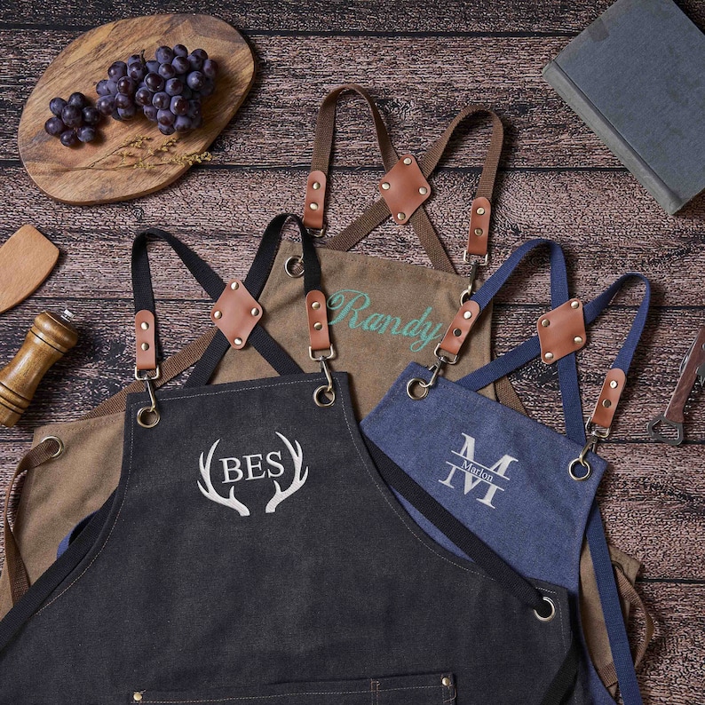 May include: Three personalized denim aprons with adjustable straps and leather accents. The aprons are embroidered with the names "Randy", "Marion", and "BES" with antlers.