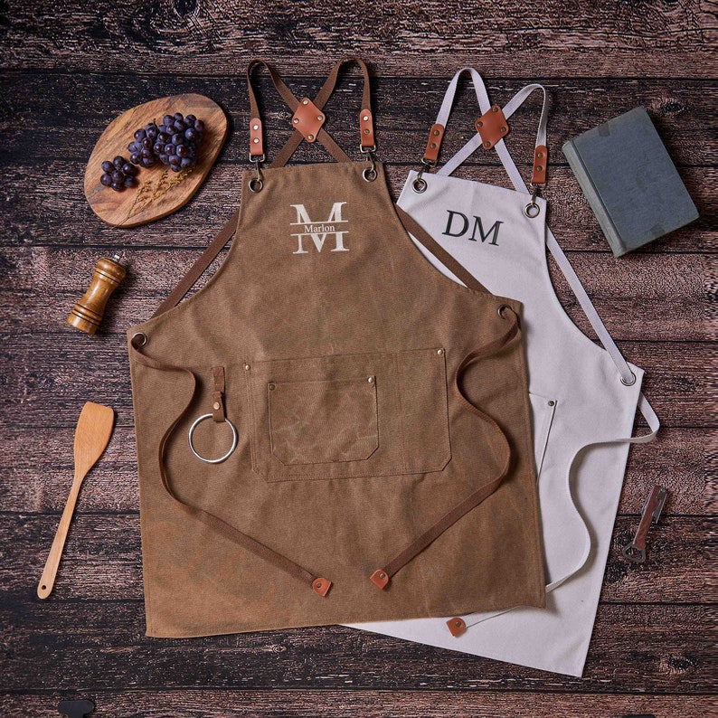 May include: Two personalized aprons, one brown with the monogram "M Marlon" and one white with the monogram "DM". Both aprons have adjustable straps and a pocket.