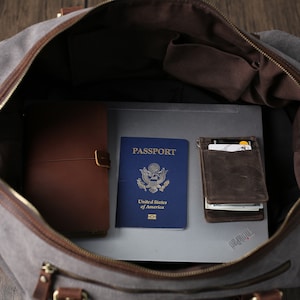 May include: A gray canvas duffel bag with brown leather accents is open and filled with travel essentials. The bag contains a passport, a leather wallet, a notebook, and a laptop.