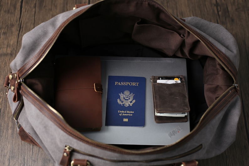 May include: A gray canvas duffel bag with brown leather accents is open and filled with travel essentials. The bag contains a passport, a leather wallet, a notebook, and a laptop.