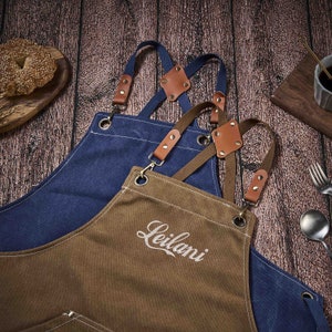 May include: Two brown canvas aprons with adjustable straps and leather accents. The apron on top has the word "Leilani" embroidered on the front.