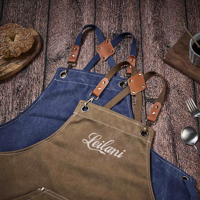 May include: Two brown canvas aprons with adjustable straps and leather accents. The apron on top has the word "Leilani" embroidered on the front.