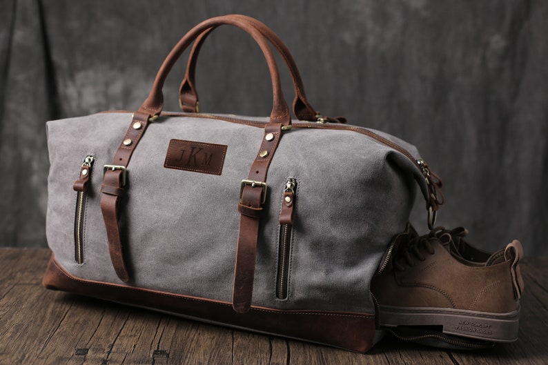 May include: A gray canvas duffel bag with brown leather accents and a personalized leather patch with the initials "J.K.M.". The bag has two side pockets with zippers and a top handle.