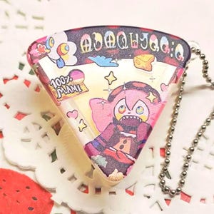 May include: A triangular keychain with a cartoon character design. The keychain features a pink and brown character with a star-shaped head. The design includes the text "100% MANI" and other colorful illustrations. A silver ball chain is attached.