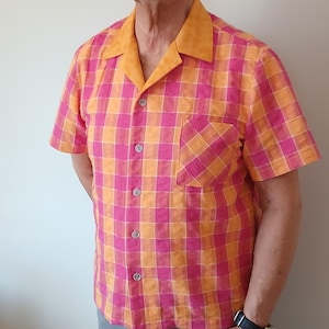 May include: Short-sleeved shirt with a vibrant plaid pattern in shades of pink and orange. The shirt features a contrasting orange collar and a single chest pocket. It has a button-down front and short sleeves, offering a casual style.