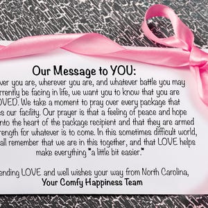 Pu&ograve; includere: Un biglietto bianco con un nastro rosa e il testo "Our Message to YOU:" seguito da un messaggio d'amore e sostegno. Il biglietto &egrave; su una superficie strutturata grigia e nera.
